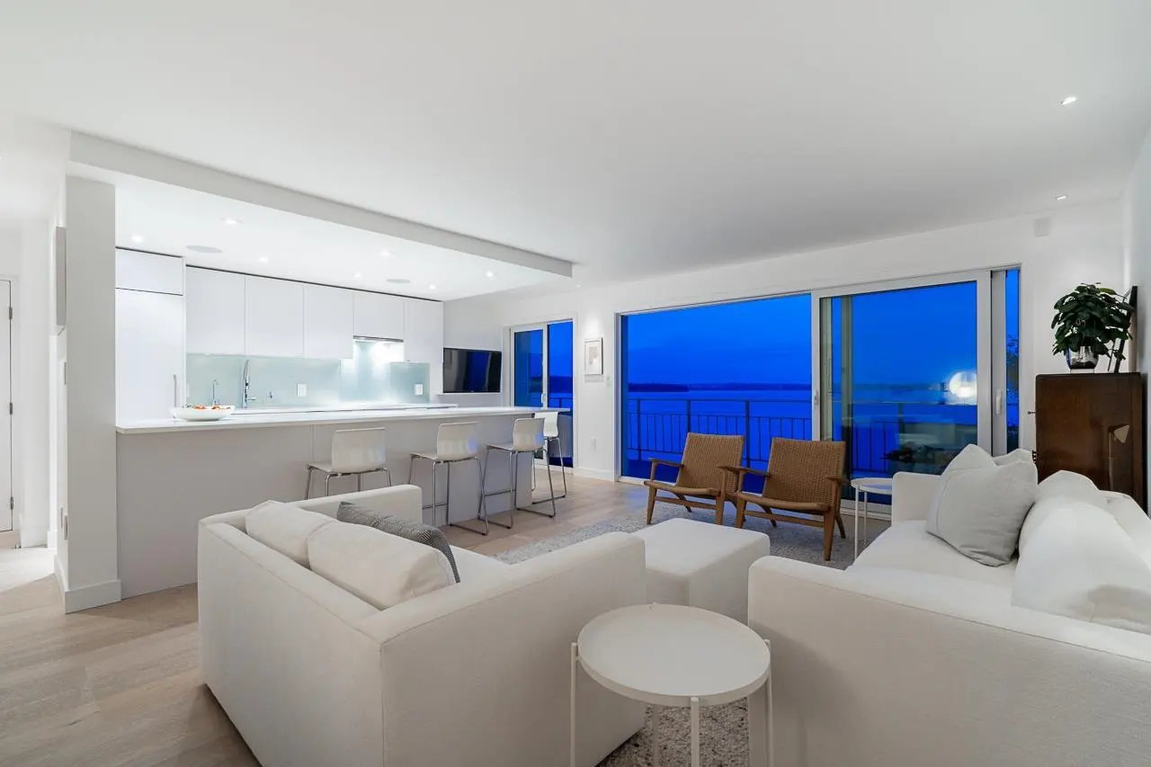 Bright, modern living room with white sofas, kitchen bar, and ocean view through large windows at dusk.