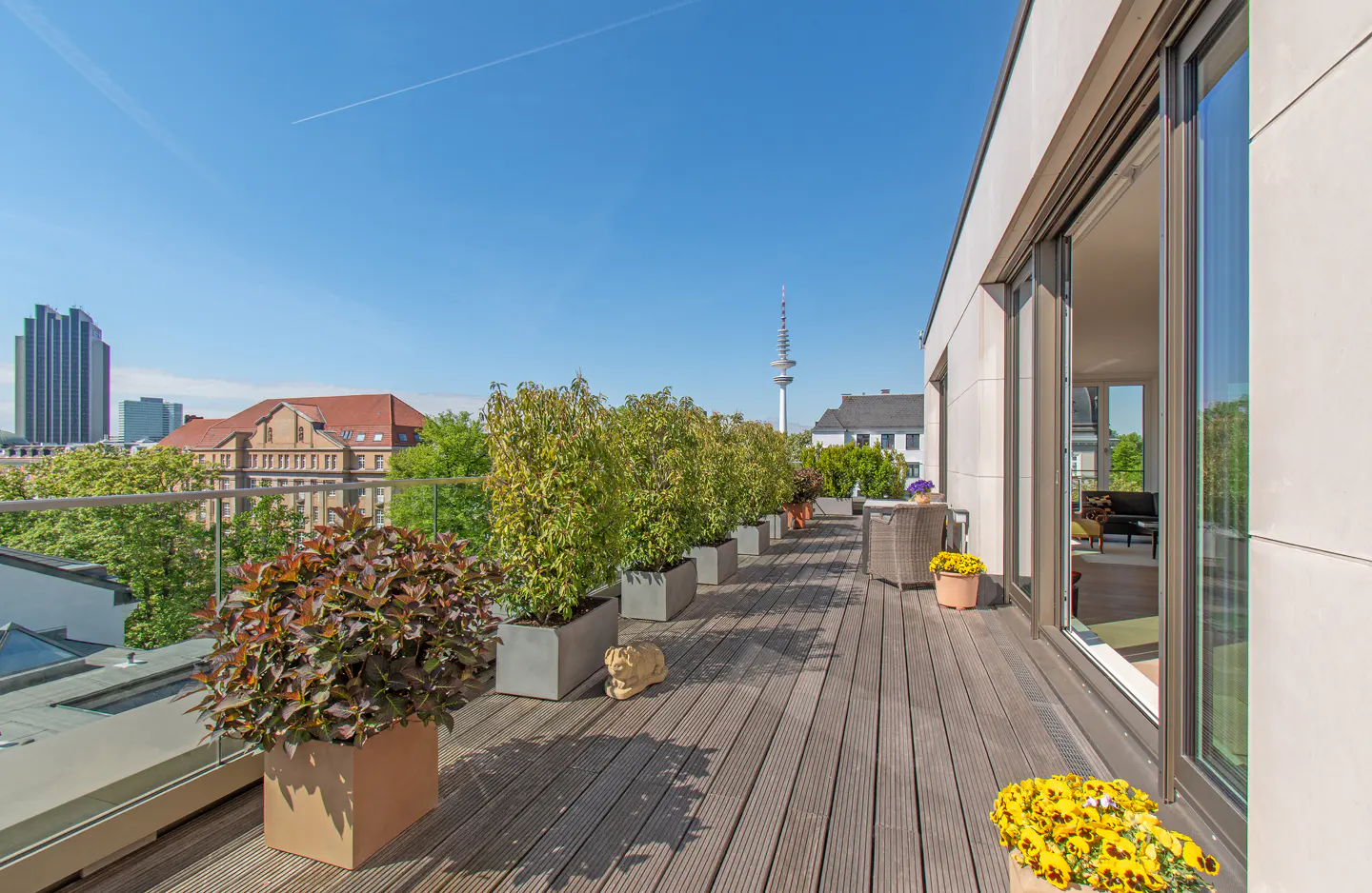 Wide balcony with wood floor, potted plants, and city view. Sliding glass doors lead inside. Blue sky above.