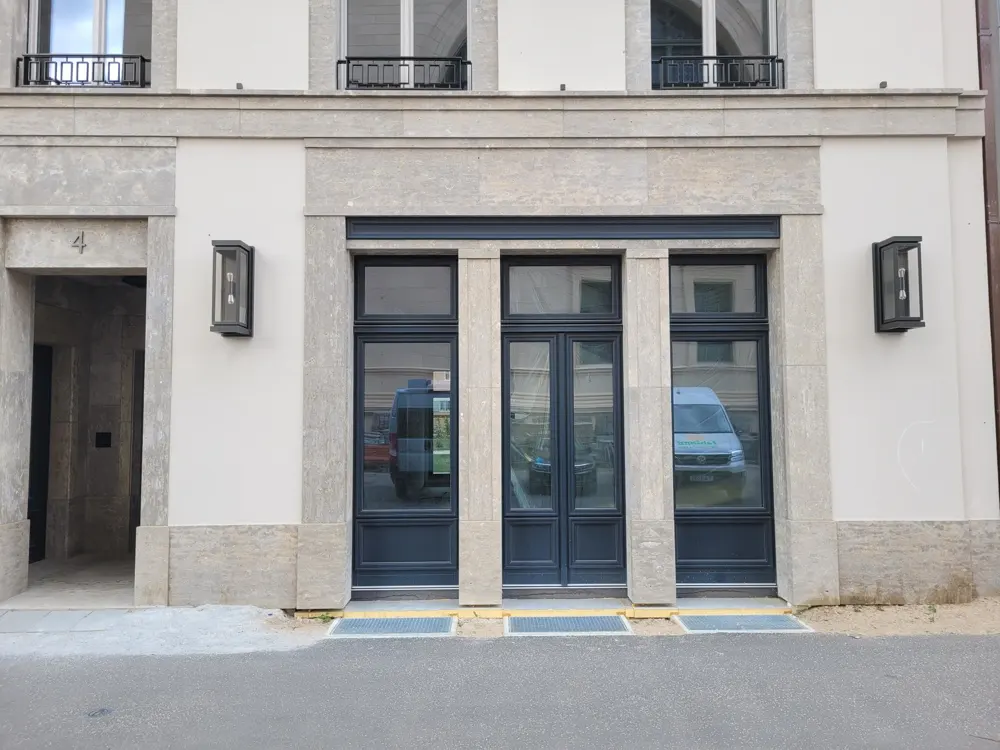 Exterior view of a modern building with a beige stone facade, dark grey double doors, and matching window frames.