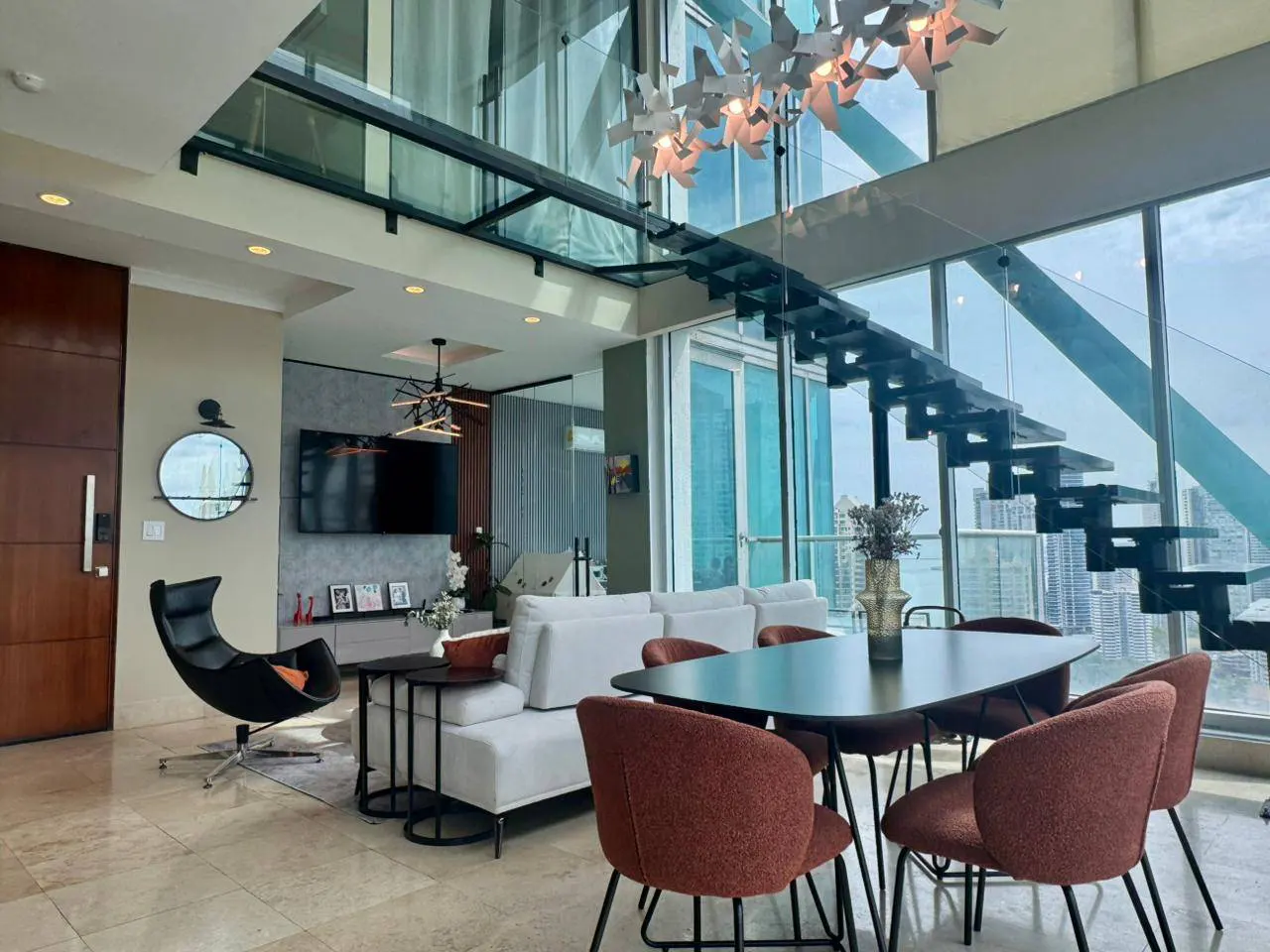 Modern apartment interior with a black staircase, white sofa, dining table with rust-colored chairs, and city views through large windows.