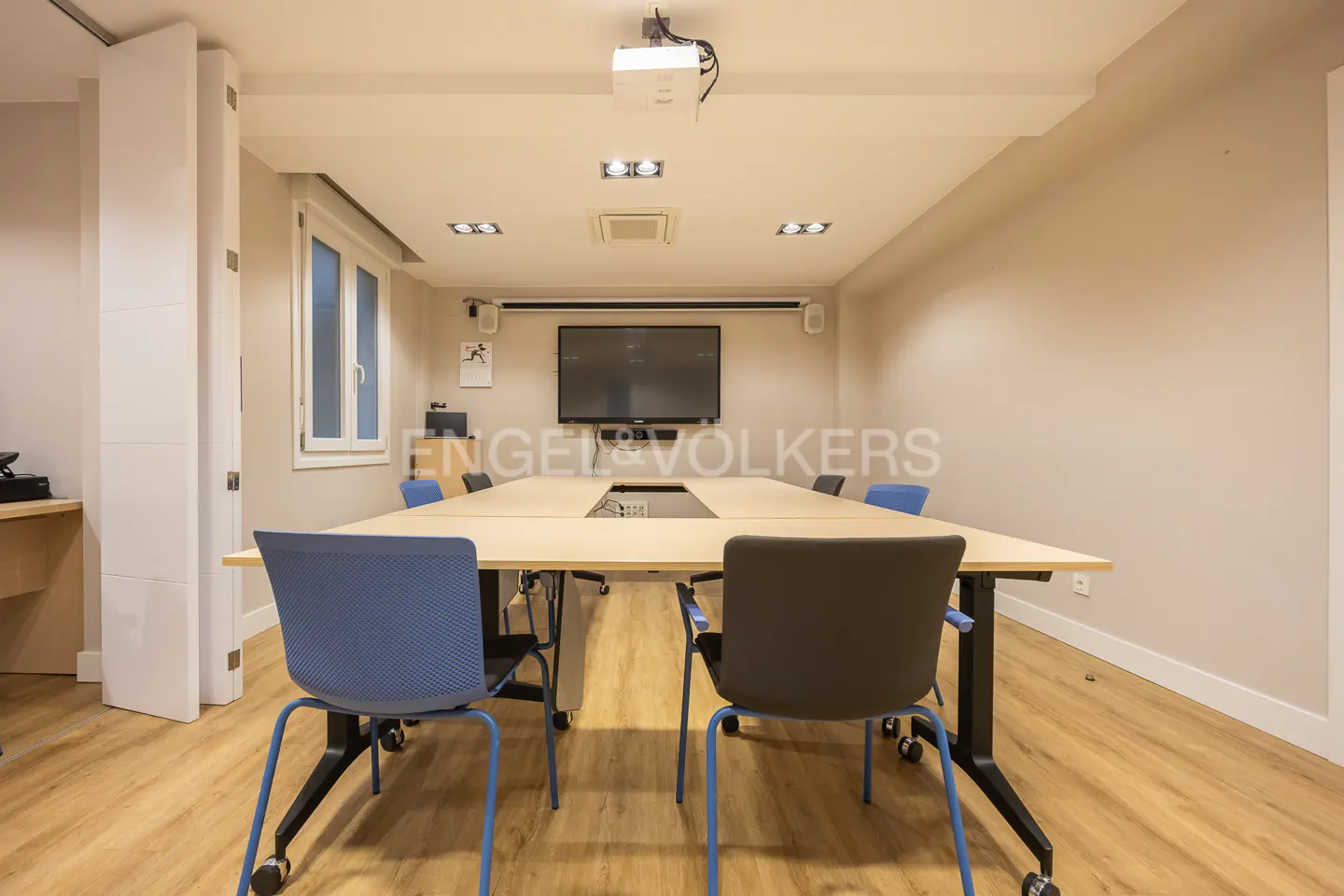 A conference room with a large table, blue and gray chairs, a projector, and a TV screen. The room has wood floors and beige walls.