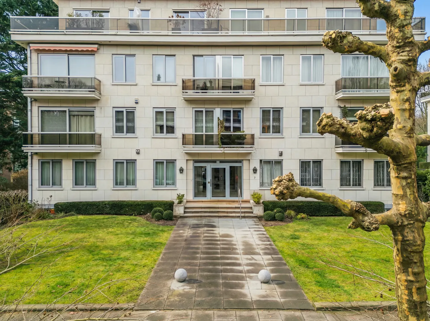 Four-story apartment building with balconies, a stone walkway, and green grass. A tree is on the right.