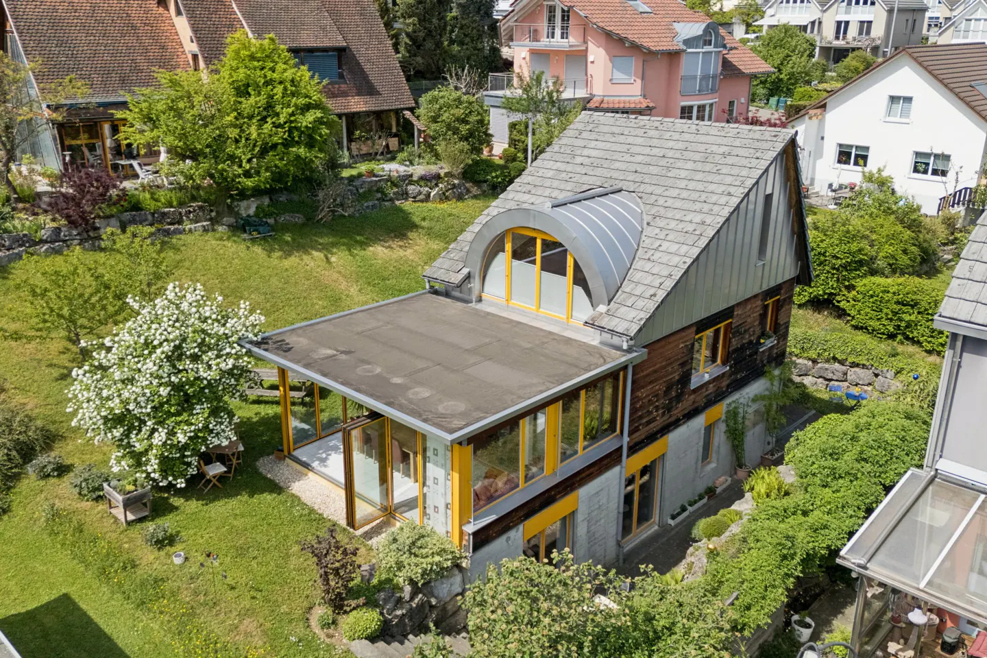 Aerial view of a modern house with yellow window frames, a flat roof extension, and a gray shingled roof, surrounded by lush greenery.