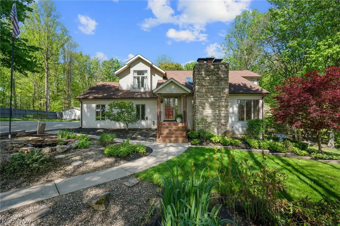 Two-story house with stone chimney, brown roof, and beige siding. A walkway leads to the front door with green trees in the background.