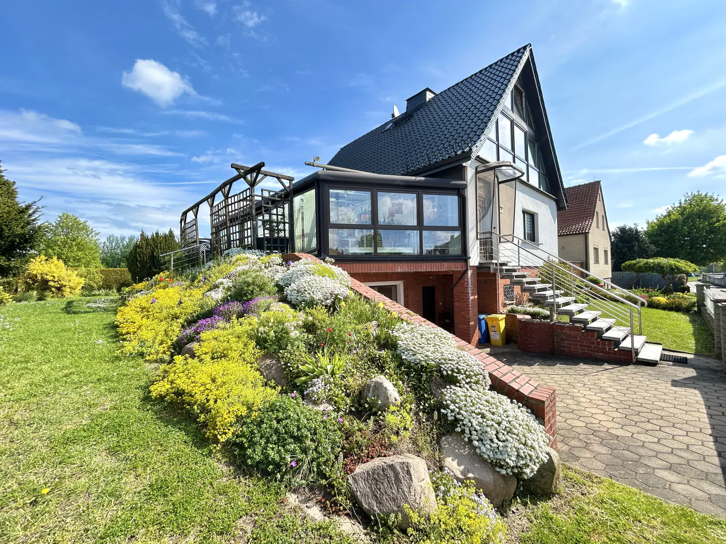 Exterior view of a two-story house with a black roof, white walls, and a glass-enclosed sunroom. A flower-covered slope leads to the house, with stairs to the right.