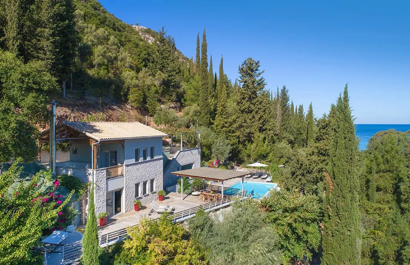 A stone villa with a pool and patio nestled in a lush, tree-covered hillside overlooking the blue sea under a clear sky.