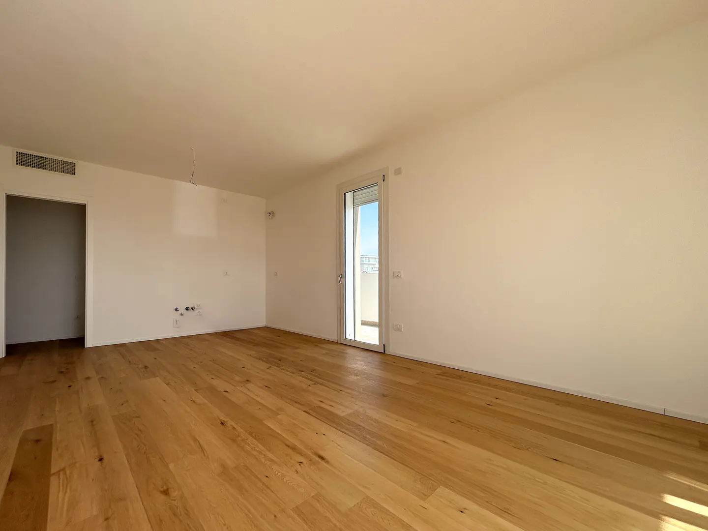Empty room with light wood floors, white walls, and a sliding glass door to a balcony with a city view.