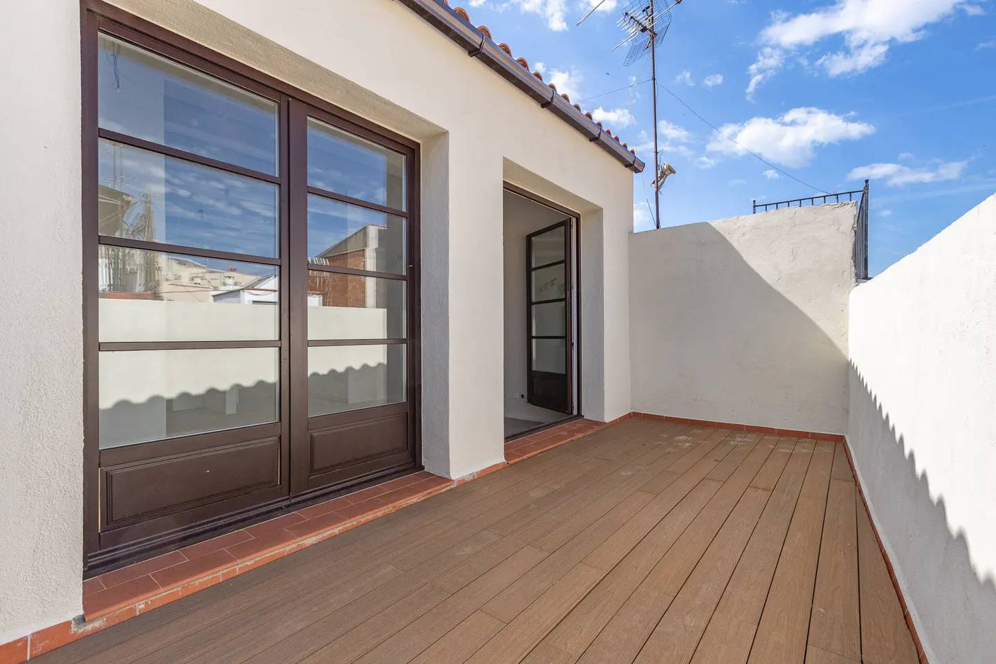 Outdoor terrace with brown wood flooring, white walls, and brown framed glass doors. Blue sky visible.