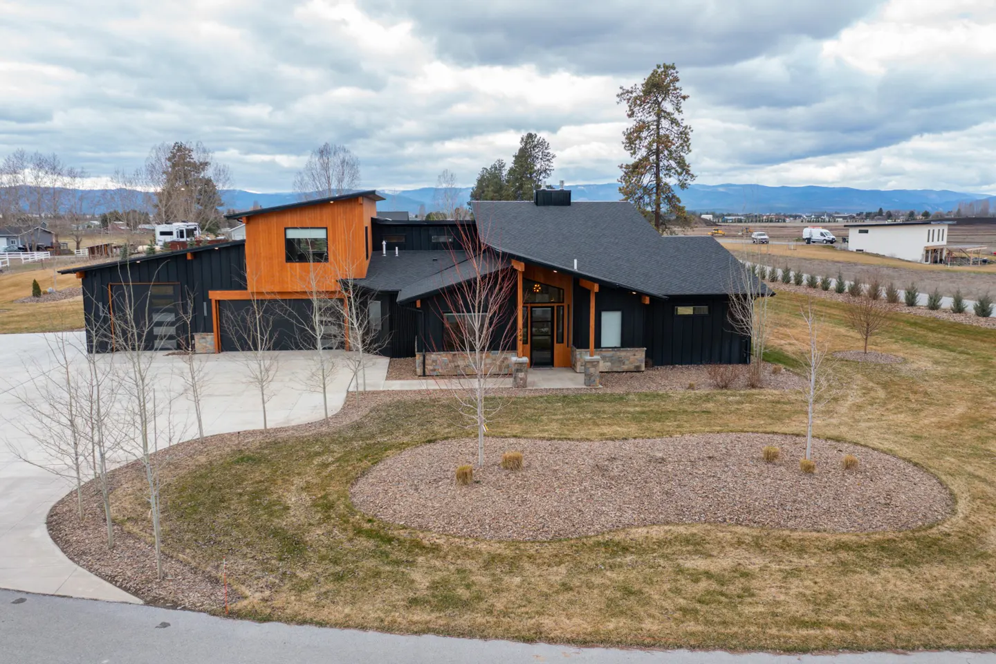 Modern black house with wood accents, a dark roof, and a circular driveway with young trees on a cloudy day.