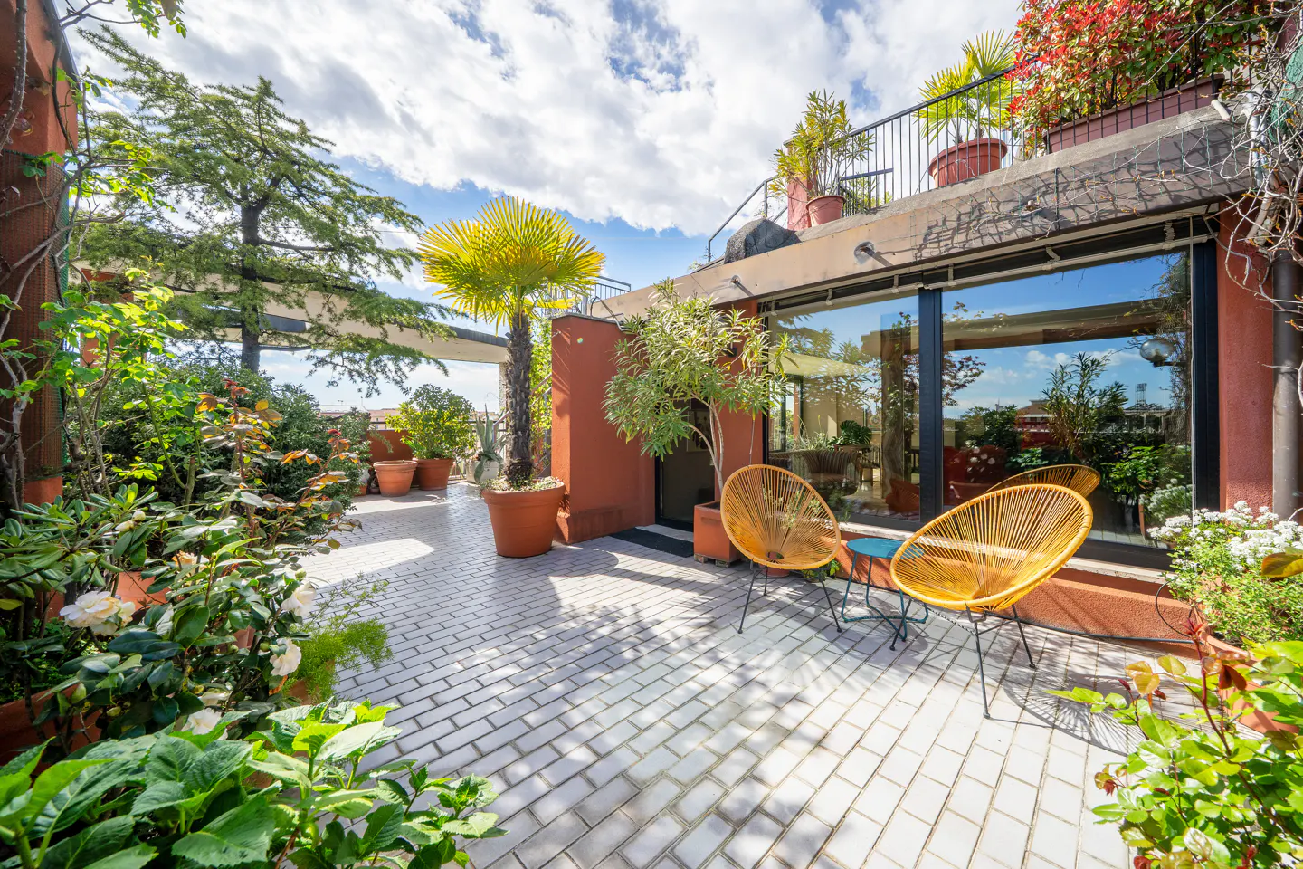 A rooftop patio with orange chairs, potted plants, and a brick floor under a partly cloudy sky.
