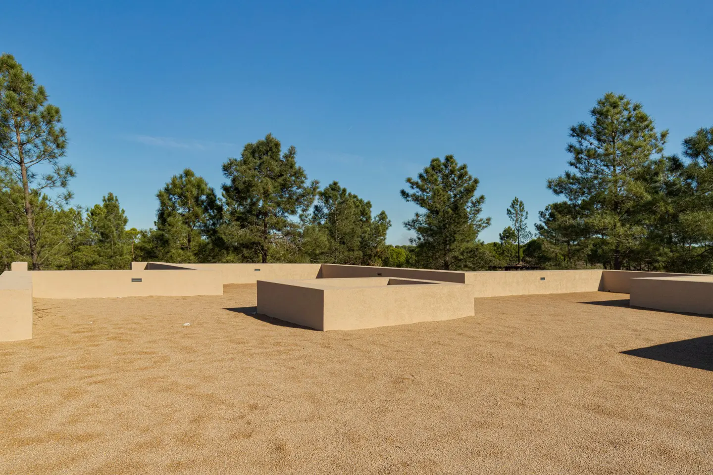 Tan gravel rooftop with low walls and cutouts, against a backdrop of green trees and a clear blue sky.