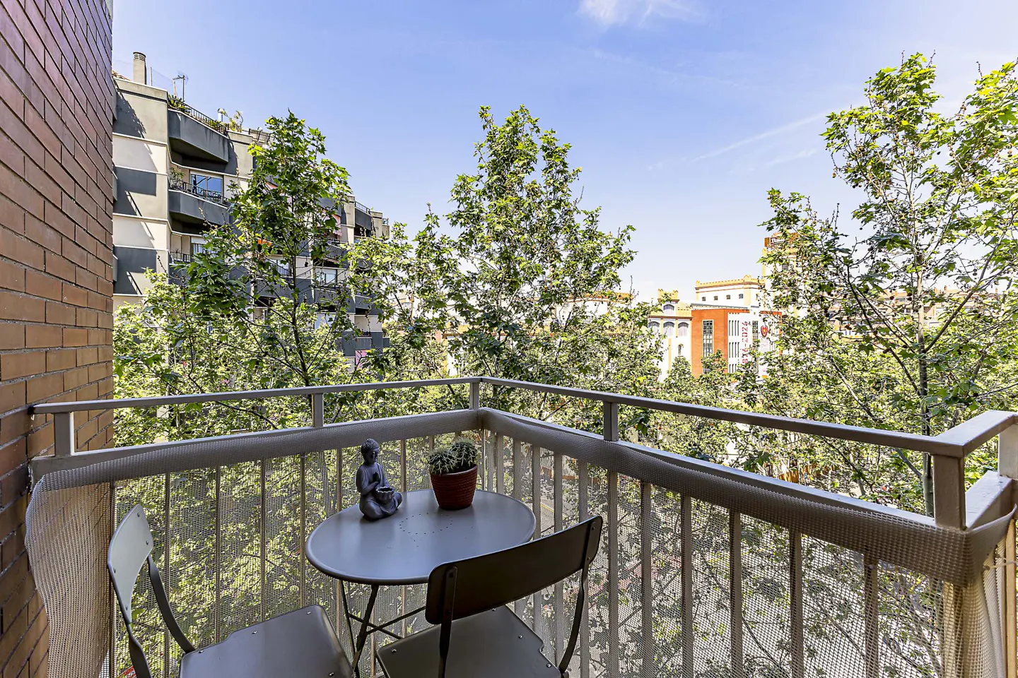 Balcony with a small round table, two chairs, a Buddha statue, and a potted plant overlooking trees and buildings.