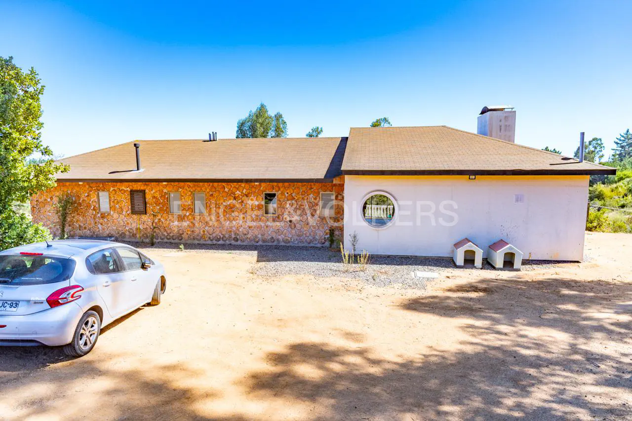 Exterior view of a one-story house with stone and white walls, a brown roof, and a white car parked nearby. Two small dog houses are visible.