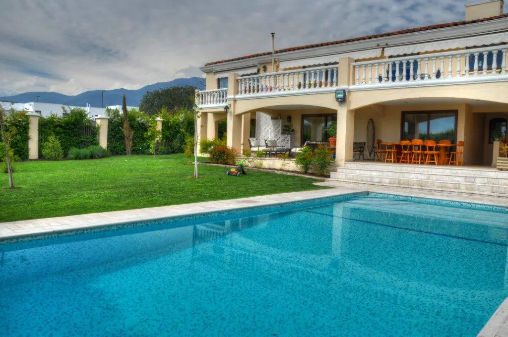 A large blue swimming pool in front of a beige two-story house with a green lawn.