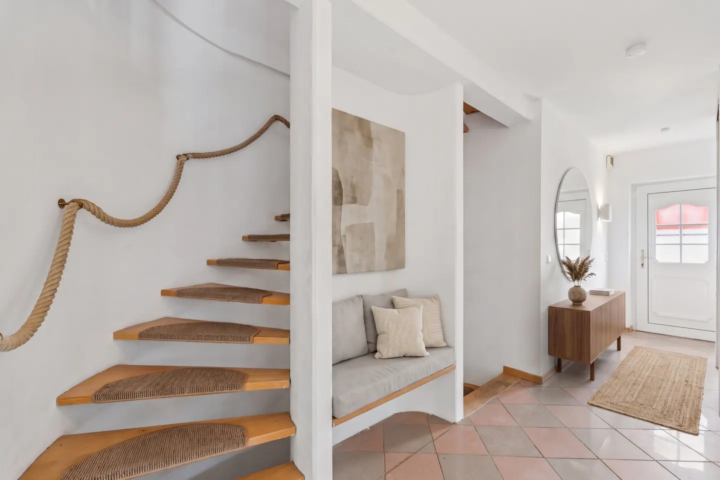 Bright entryway with a winding staircase, rope railing, and a built-in bench with pillows. A wooden cabinet and round mirror accent the space.