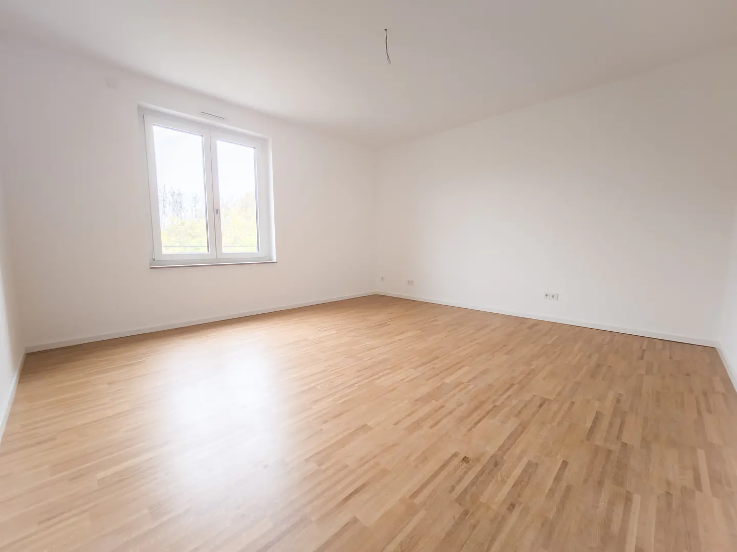 Empty room with light wood floors and white walls. A window lets in natural light.