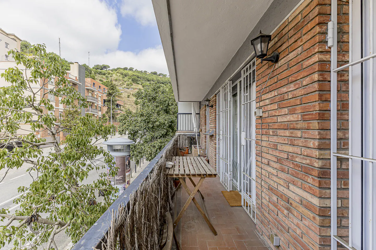 Balcony view with a wooden table, brick wall, and metal railing. Trees and buildings are visible in the background.