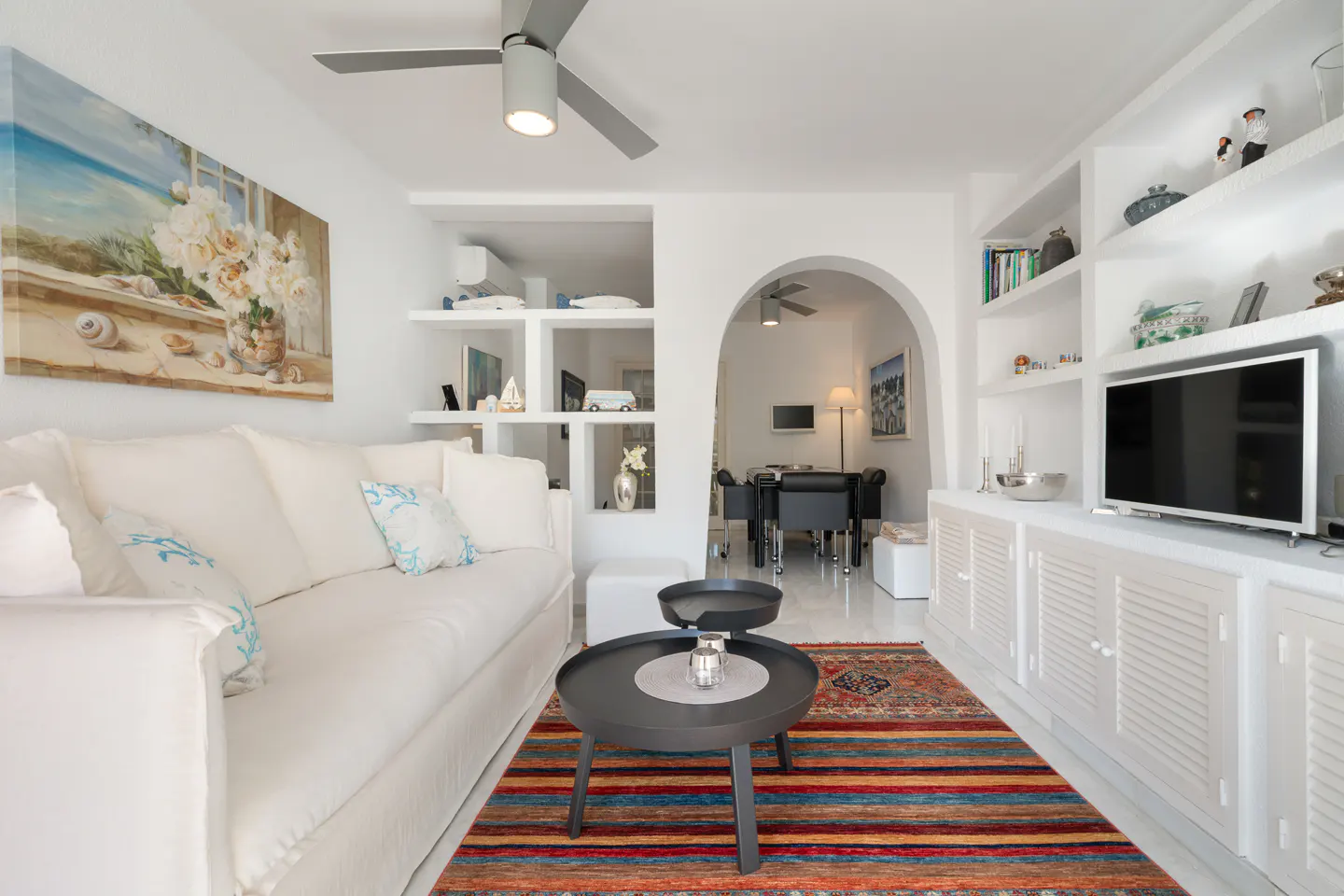 Bright living room with white sofa, gray tables, and colorful rug. A painting and built-in shelves decorate the white walls.