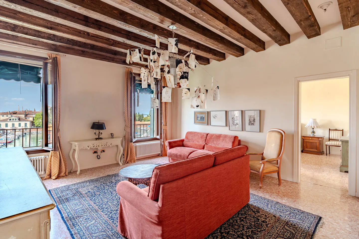Living room with red sofas, a blue rug, and a view of Venice through open windows. A unique chandelier hangs from a beamed ceiling.
