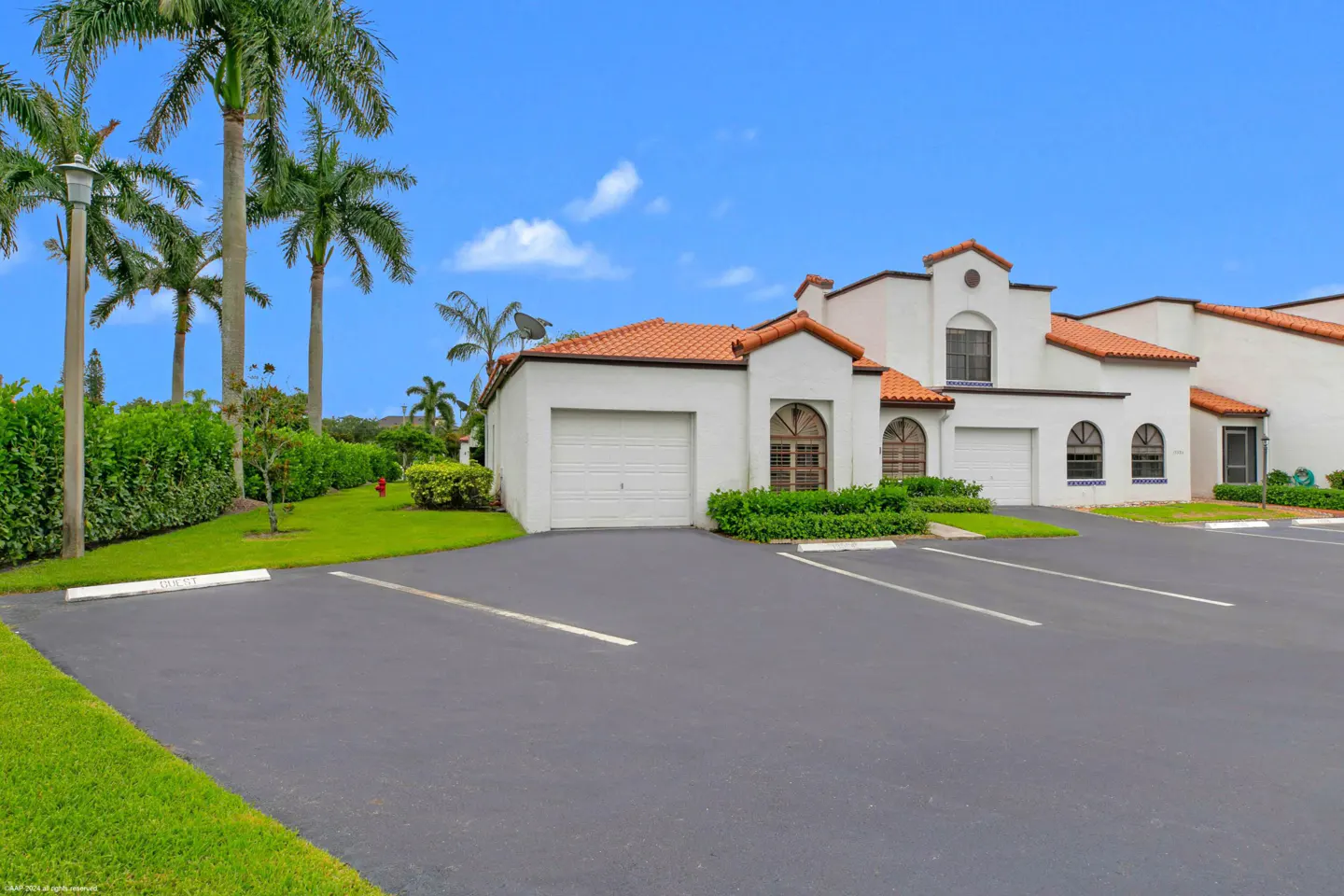 Exterior of a white stucco building with a red tile roof, palm trees, and a parking lot under a blue sky.