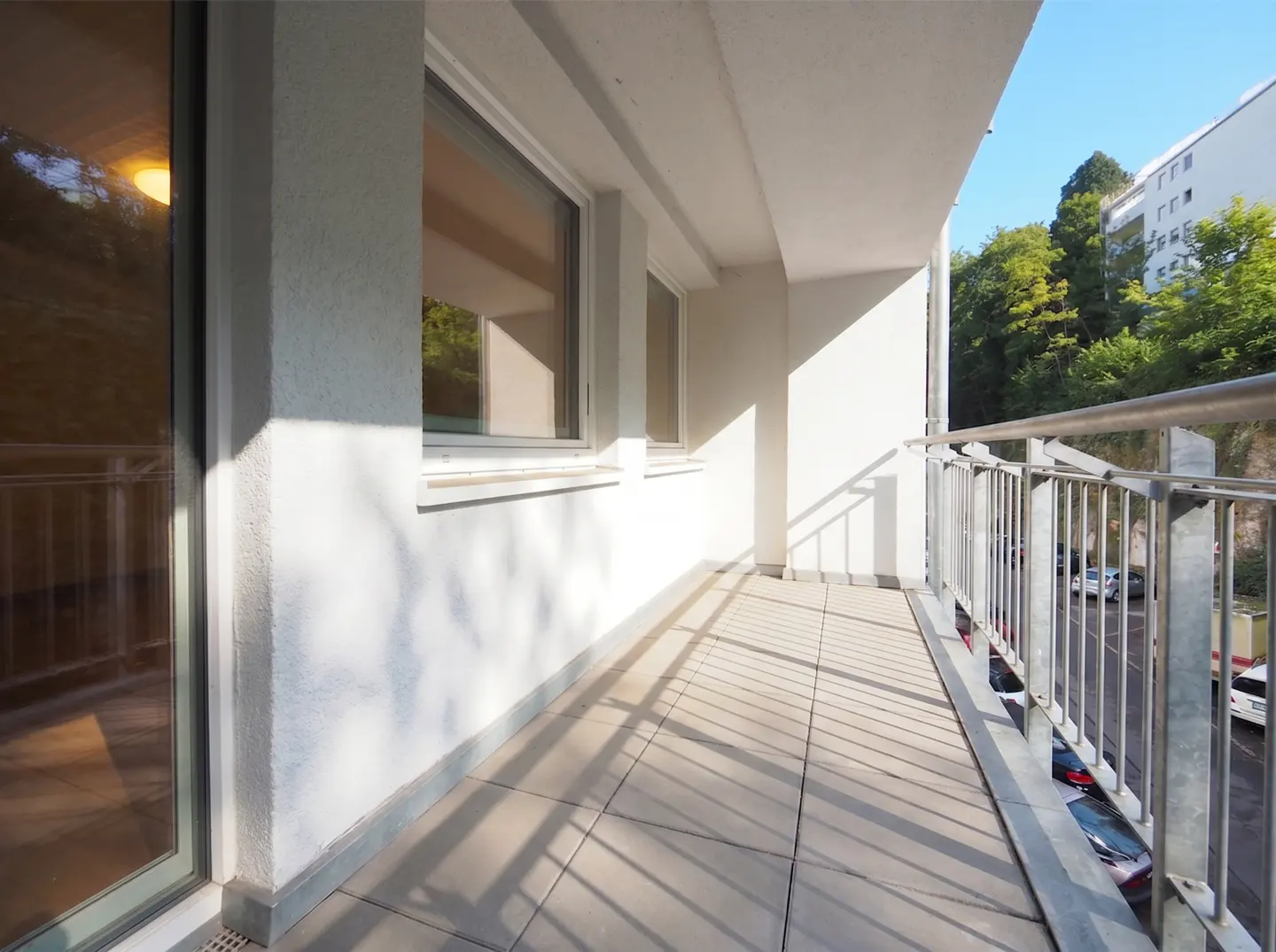A bright, empty balcony with gray tile flooring and a metal railing. Windows and a glass door are visible on the left. Trees and buildings are in the background.