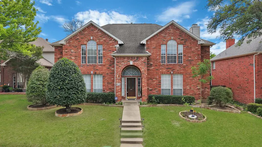 Two-story red brick house with a gray roof, arched front door, and green lawn under a blue sky.