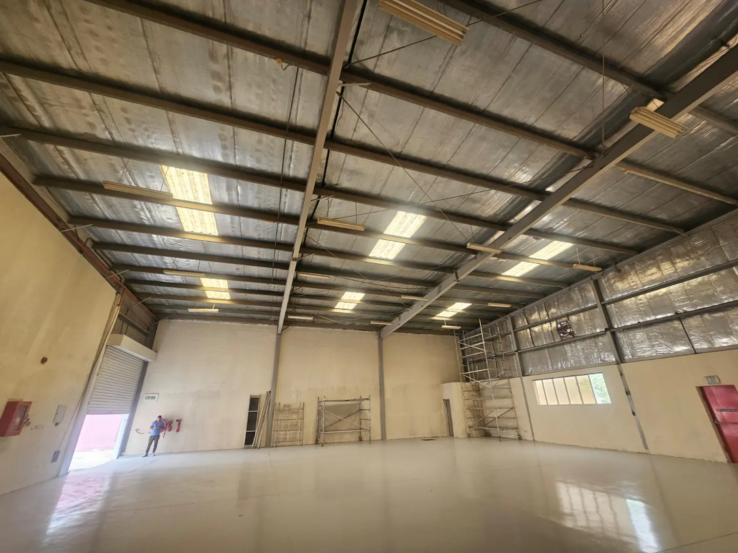 A large, empty warehouse with a high ceiling, metal beams, and a concrete floor. A man stands near the open roller door.