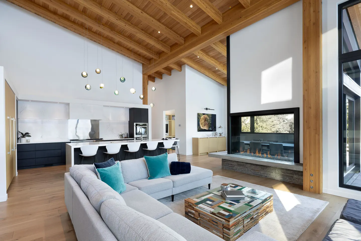 Bright, open-concept living space with wood beams, a modern fireplace, and a gray sectional sofa with teal pillows.