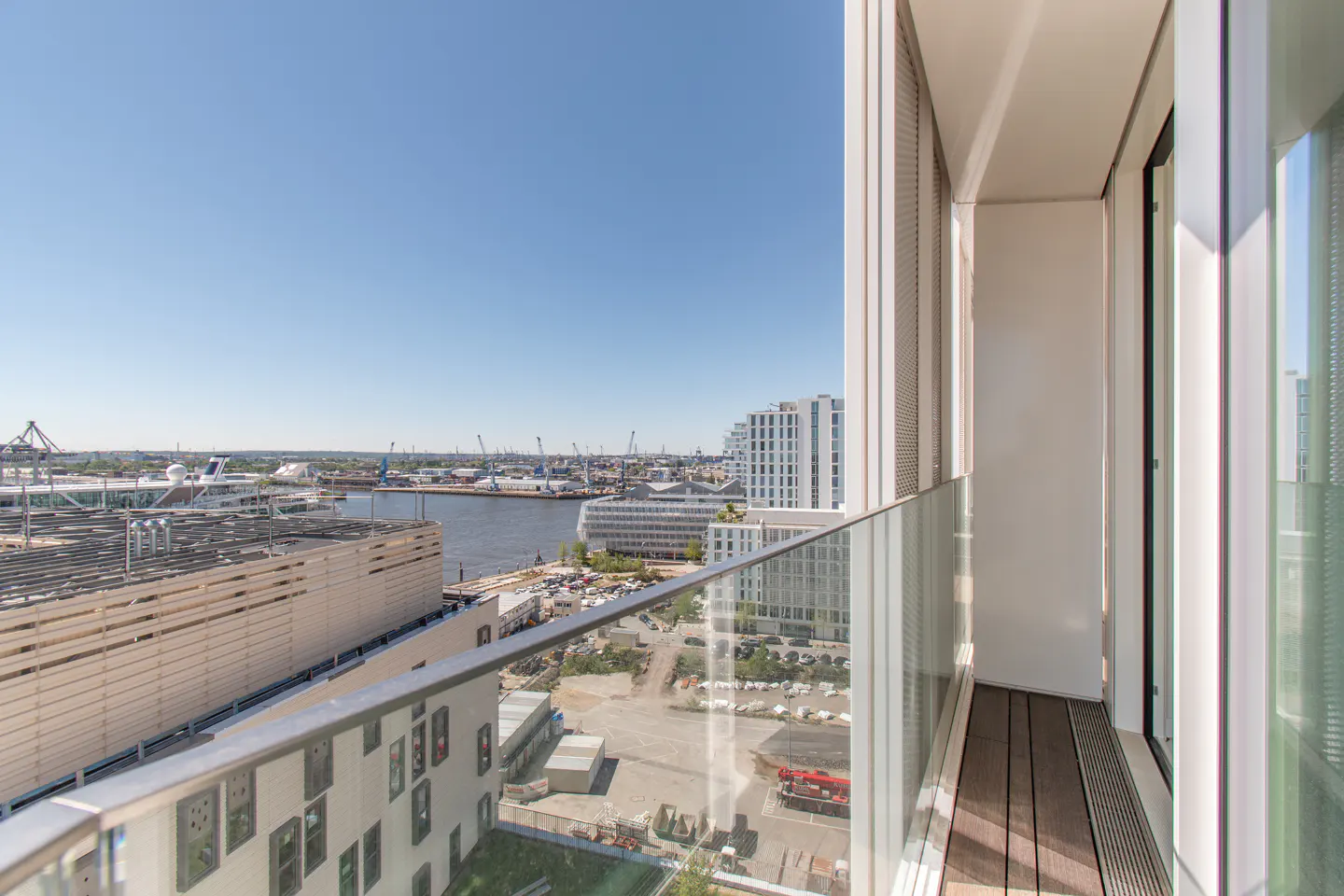 View from a modern balcony with a glass railing overlooking a harbor with cranes and buildings under a clear blue sky.