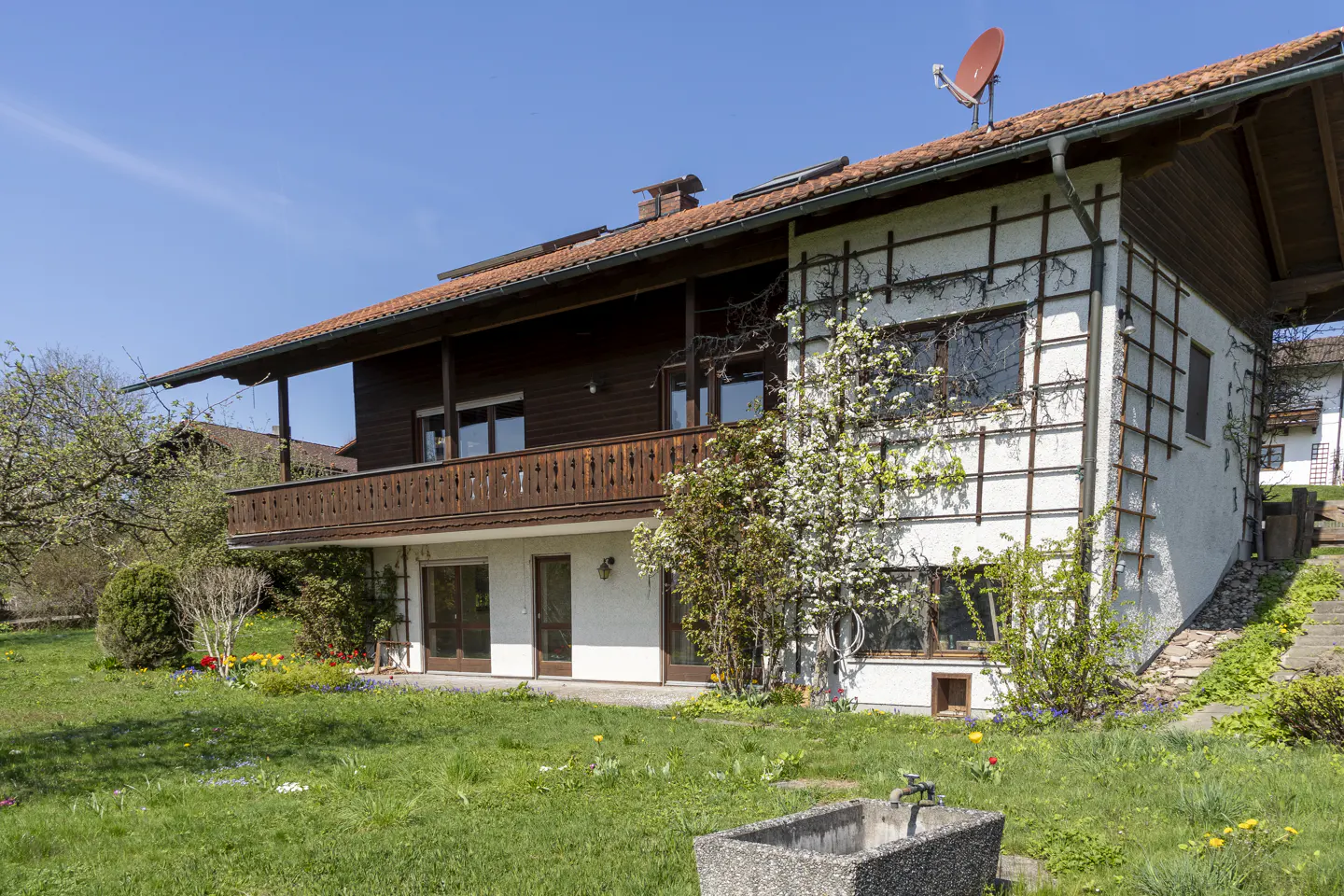 Two-story house with a brown balcony, white walls, and a red satellite dish on the roof, surrounded by a green lawn and trees.