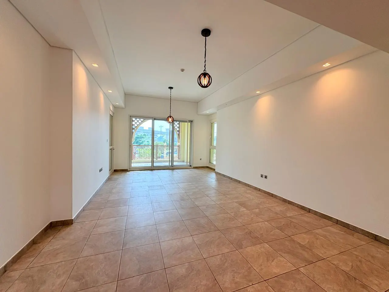 Bright, empty room with tan tile floor, white walls, and balcony doors. Two hanging lights and recessed lighting illuminate the space.