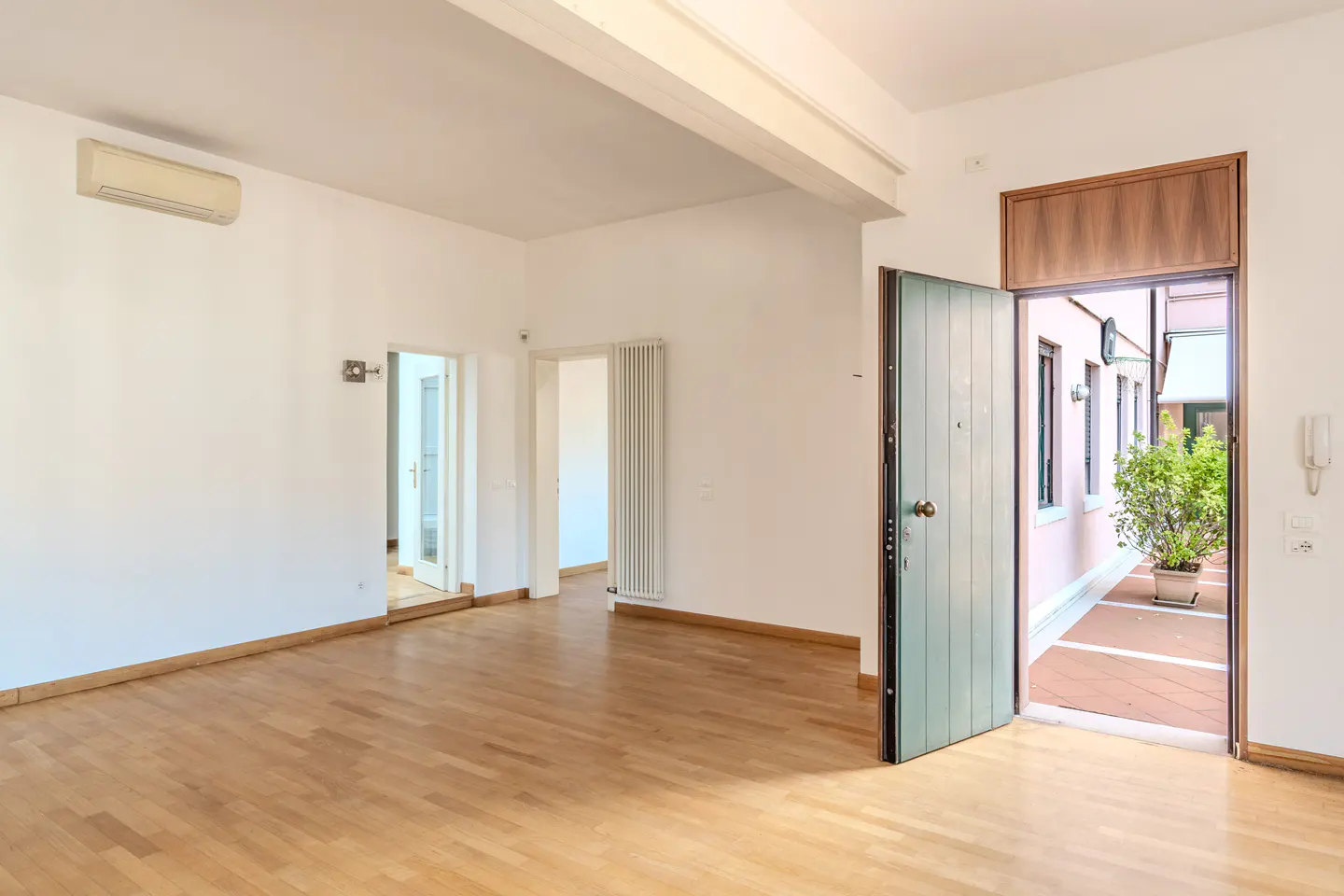 Empty room with light wood floors, white walls, an AC unit, and an open door leading to a sunny outdoor patio with plants.