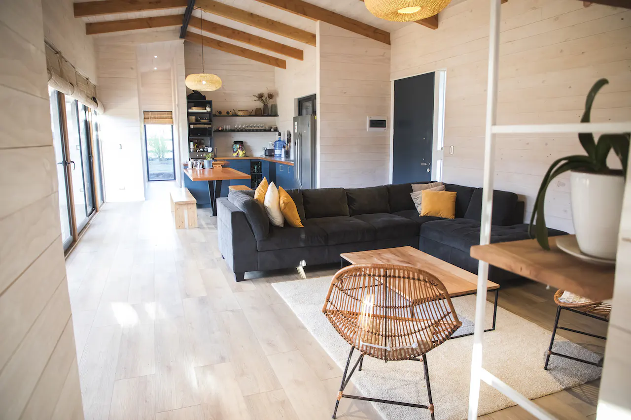 Open-plan living area with light wood walls, a dark grey sectional sofa, a rattan chair, and a blue kitchen in the back.
