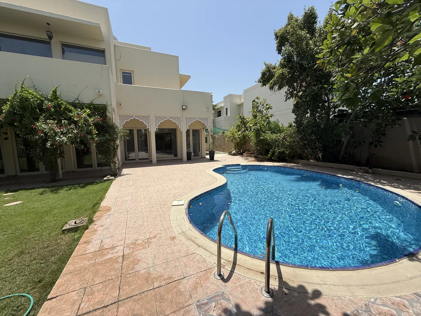 Backyard view of a two-story beige house with a blue swimming pool, brown tile patio, and green grass.