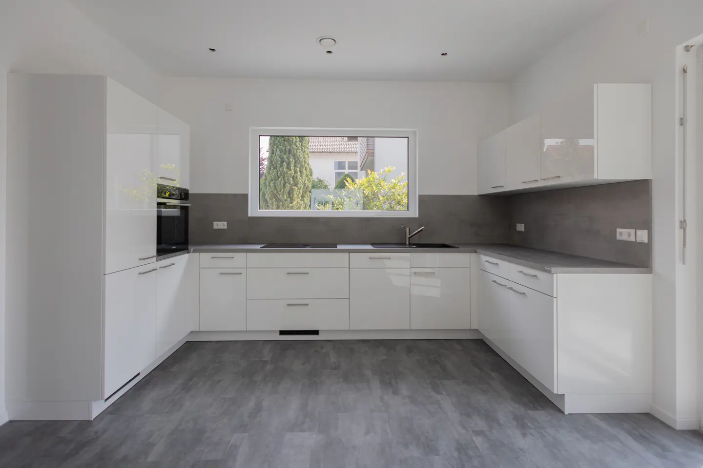 Bright, modern kitchen with white cabinets, gray countertops and flooring. A window overlooks a green yard.