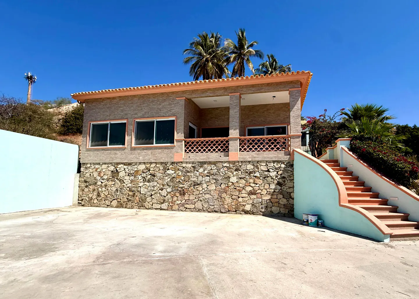 Exterior view of a tan house with a stone foundation, a balcony, and stairs with a light blue wall. Palm trees in the background.