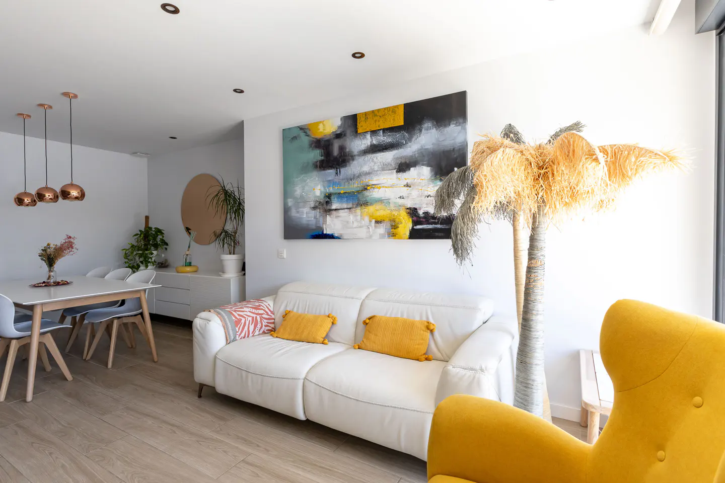 Bright living room with white walls, wood floors, and modern furniture. A white sofa with yellow pillows sits beneath an abstract painting. A dining area is visible in the background.