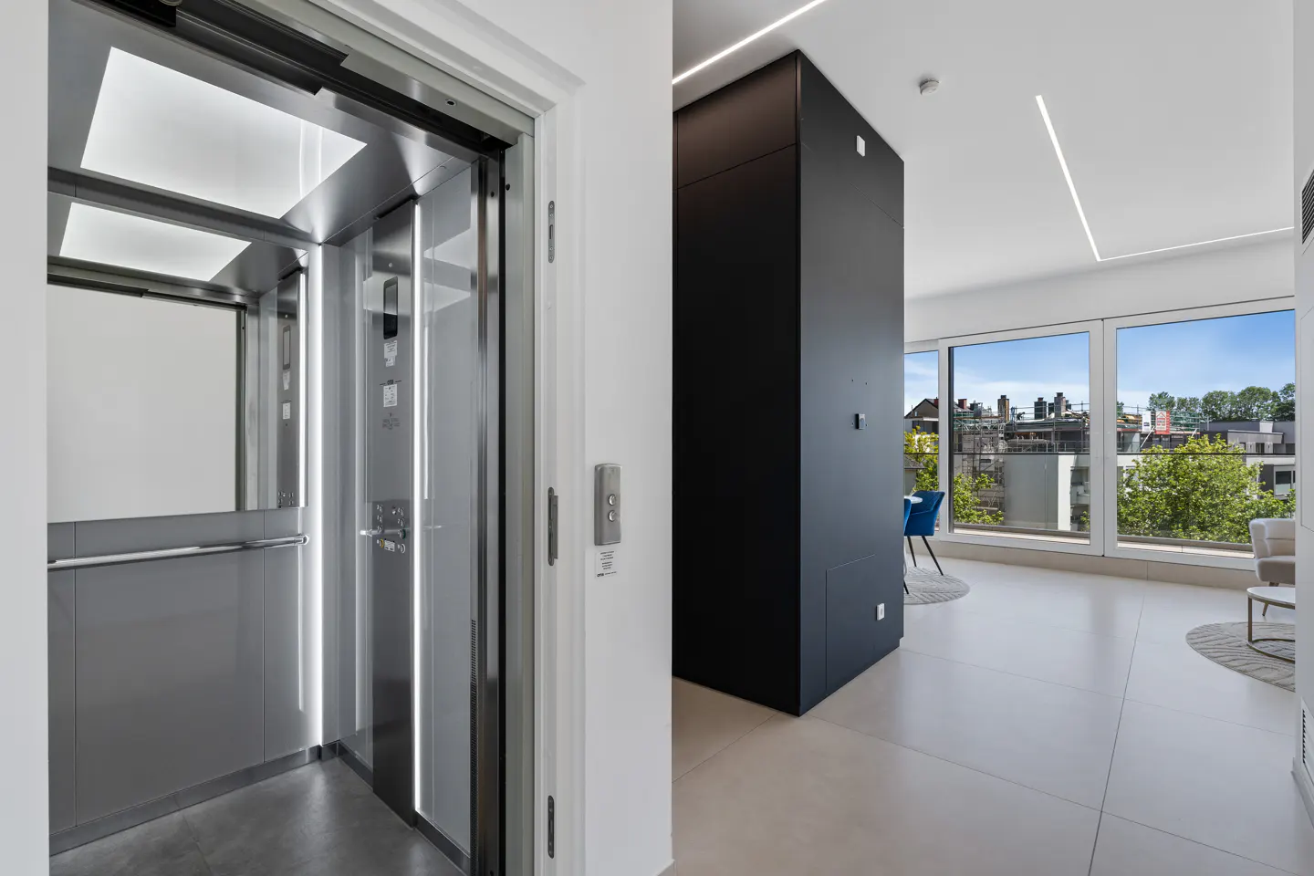 Modern apartment interior with an open elevator, a black column, and large windows overlooking a cityscape.