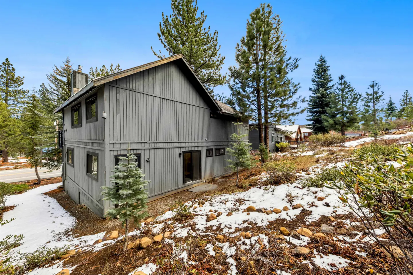 Two-story gray house with a chimney, surrounded by pine trees and snow on a sunny day.