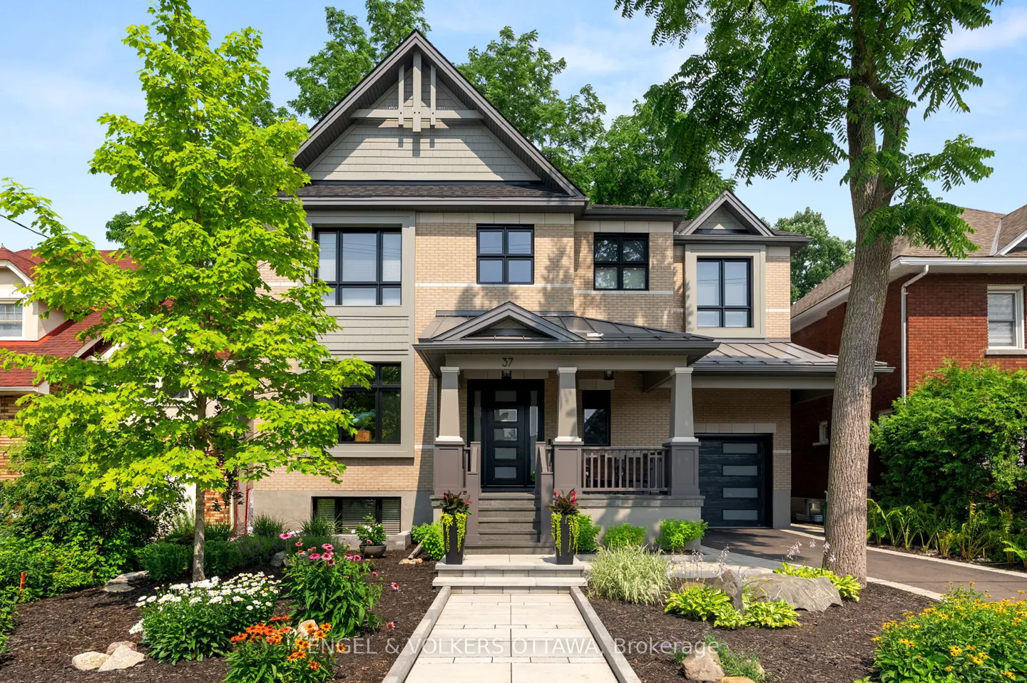 Two-story house with beige brick and gray siding, black windows, and a covered porch with gray columns. Landscaped front yard with flowers.