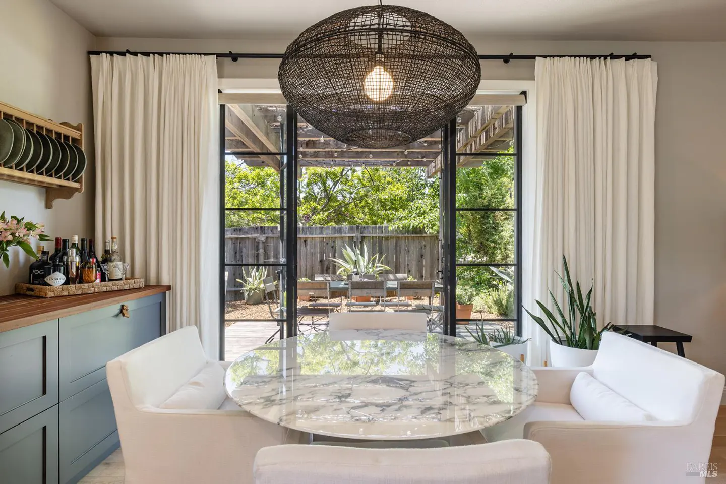 Dining room with a marble table, white chairs, and a woven pendant light. Black framed glass doors lead to a patio with greenery.