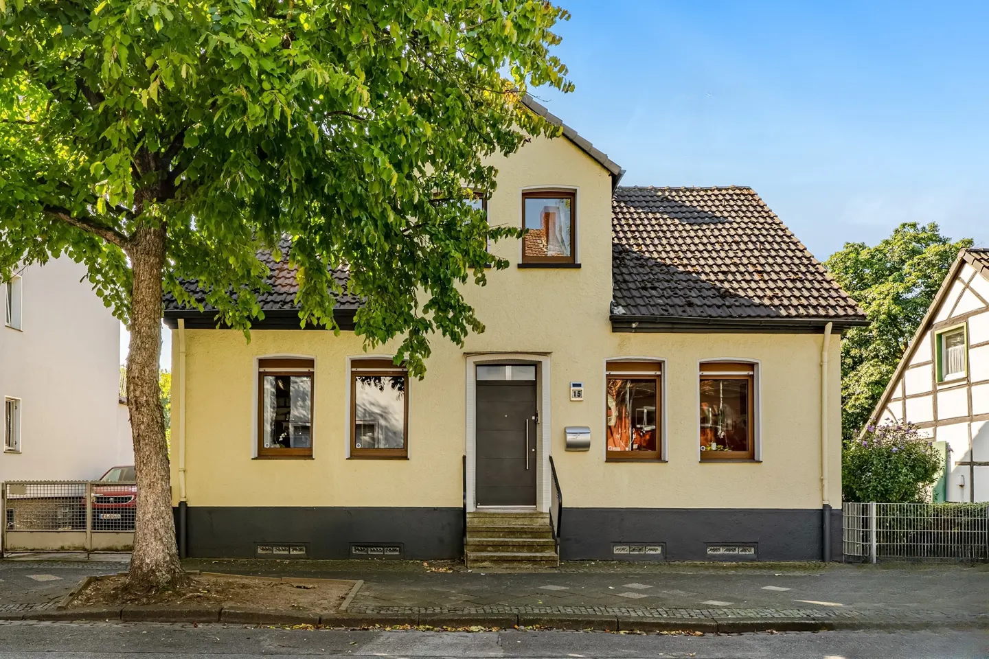Two-story yellow house with a gray door, dark roof, and a large tree on the left.