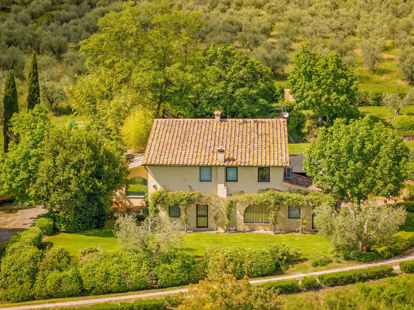Aerial view of a beige two-story house with a terracotta tile roof, surrounded by green trees and lush landscaping.