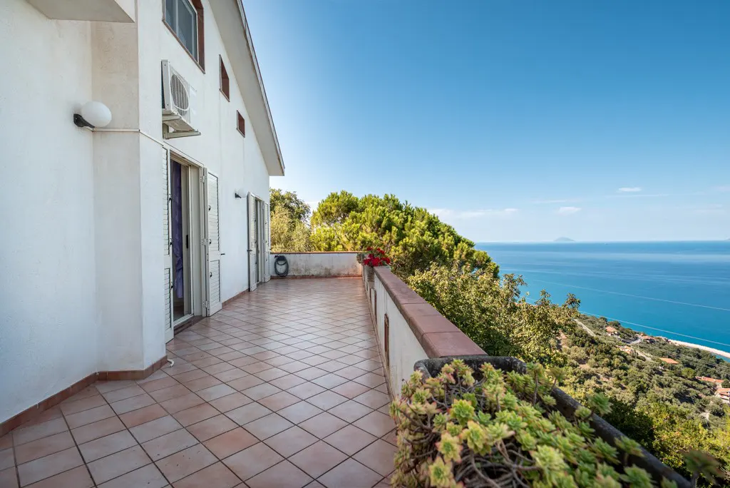 Tiled balcony of a white house overlooking a lush hillside and the blue Mediterranean sea under a clear sky.
