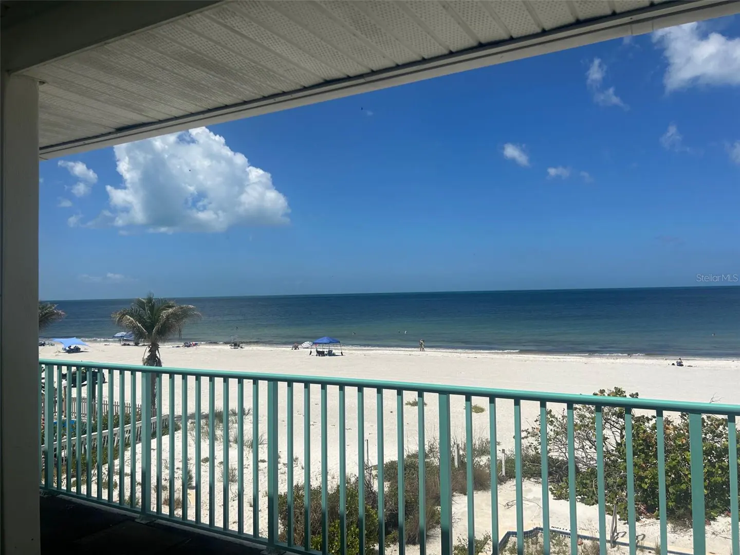 View from a balcony with a green railing overlooking a white sand beach, blue ocean, and sky with scattered clouds.