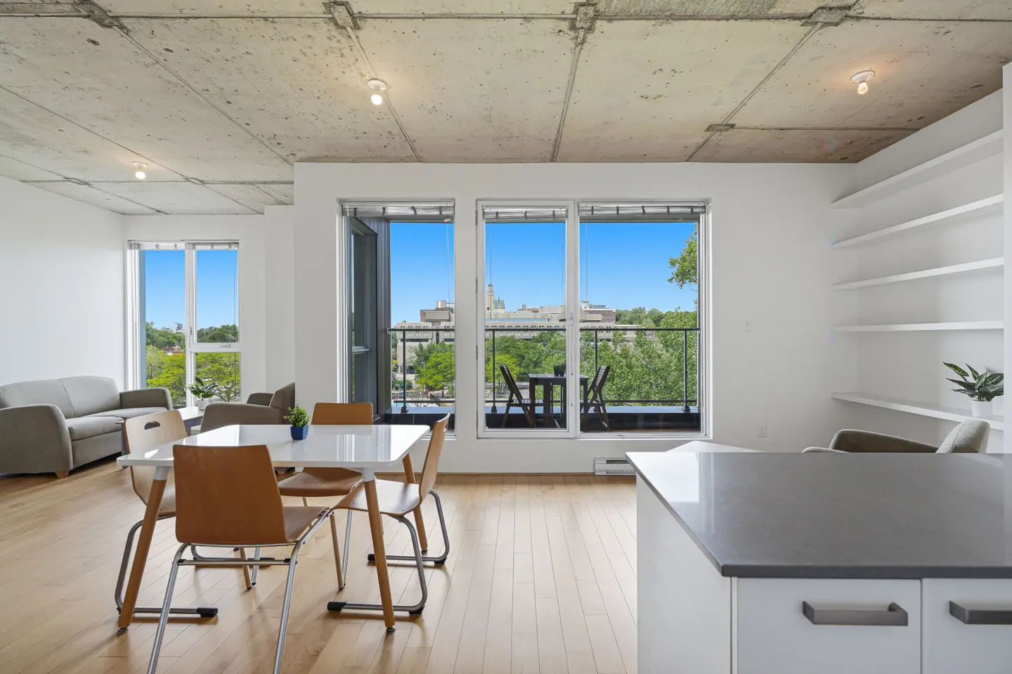 Modern apartment with concrete ceiling, dining area, white kitchen island, and balcony view of trees and city buildings.