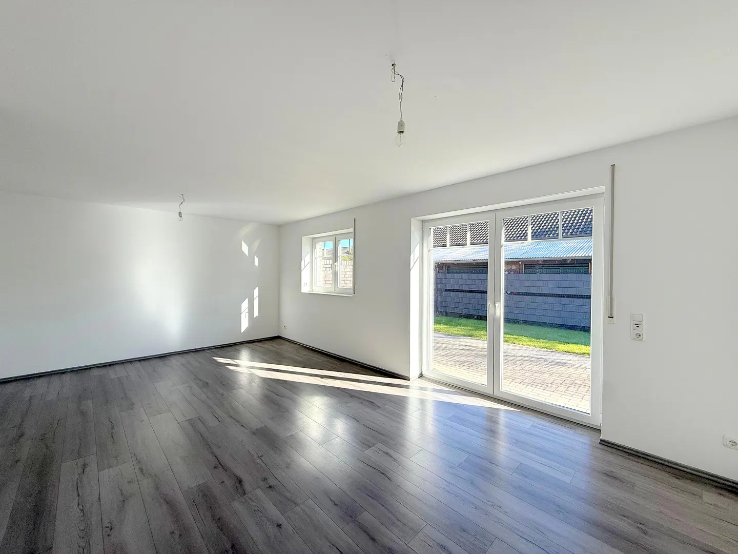 Bright, empty room with gray wood floors, white walls, and a sliding glass door to the backyard. A window is on the left wall.