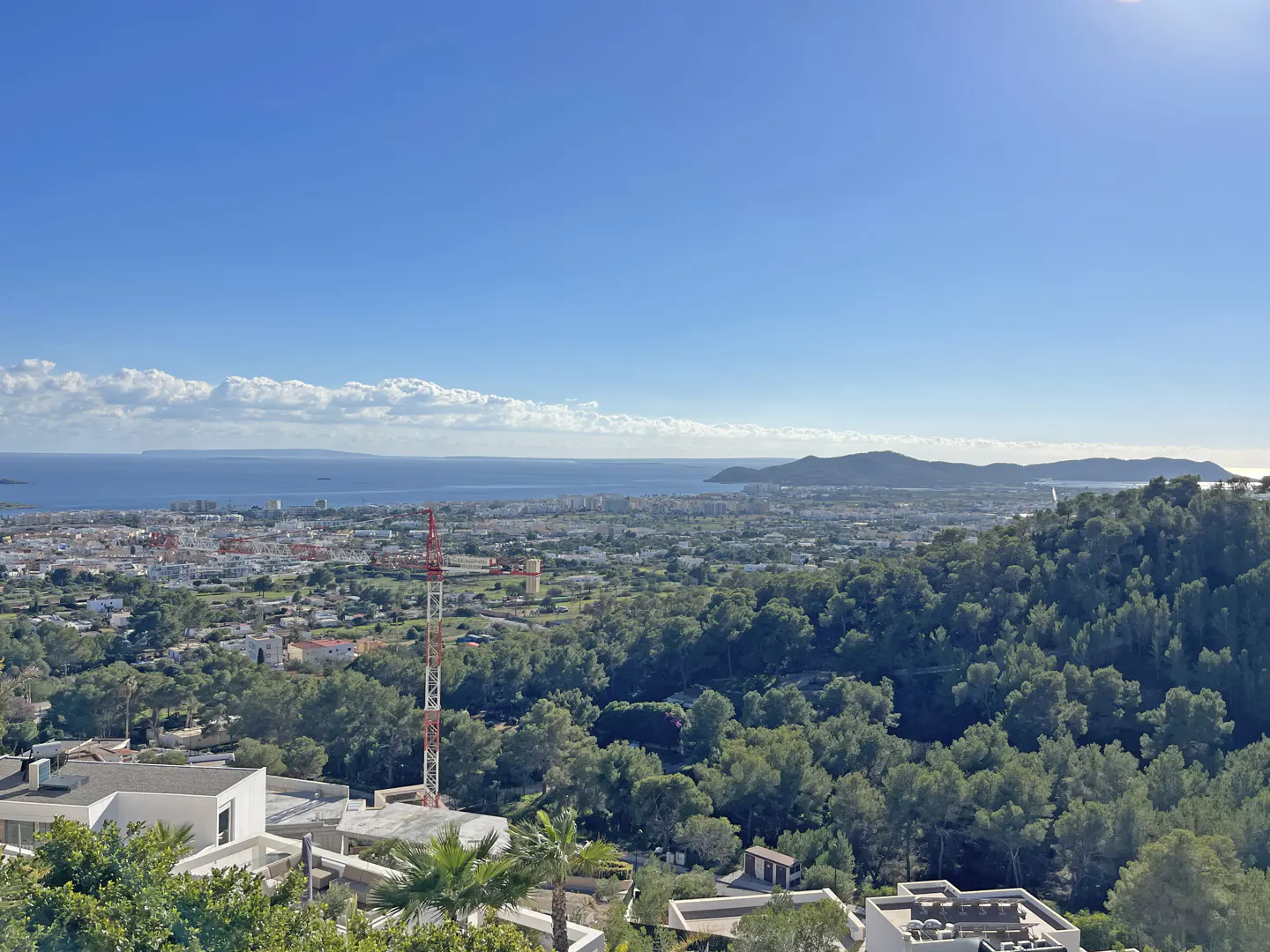 High-angle view of a coastal town with a construction crane, lush green hills, and a blue sea under a clear sky.