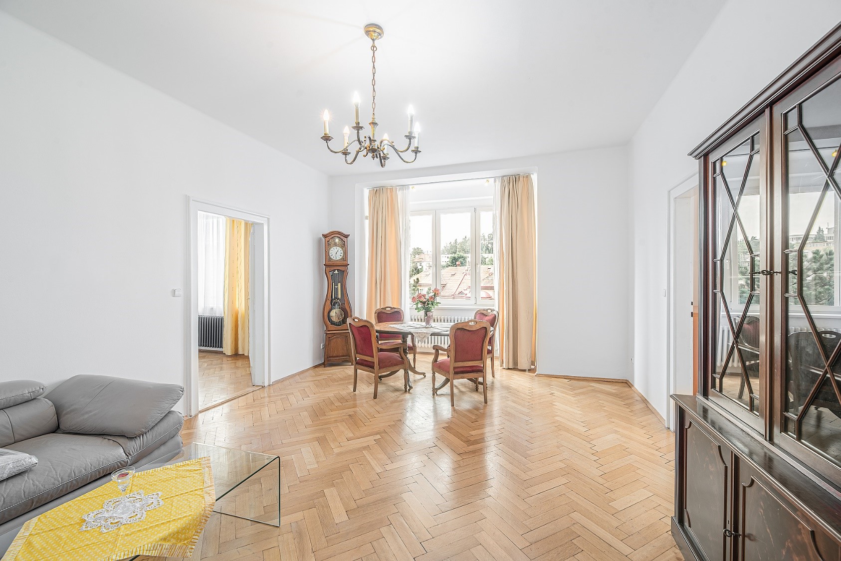 Bright dining and living room with herringbone wood floors, a chandelier, a grandfather clock, and a dining set.