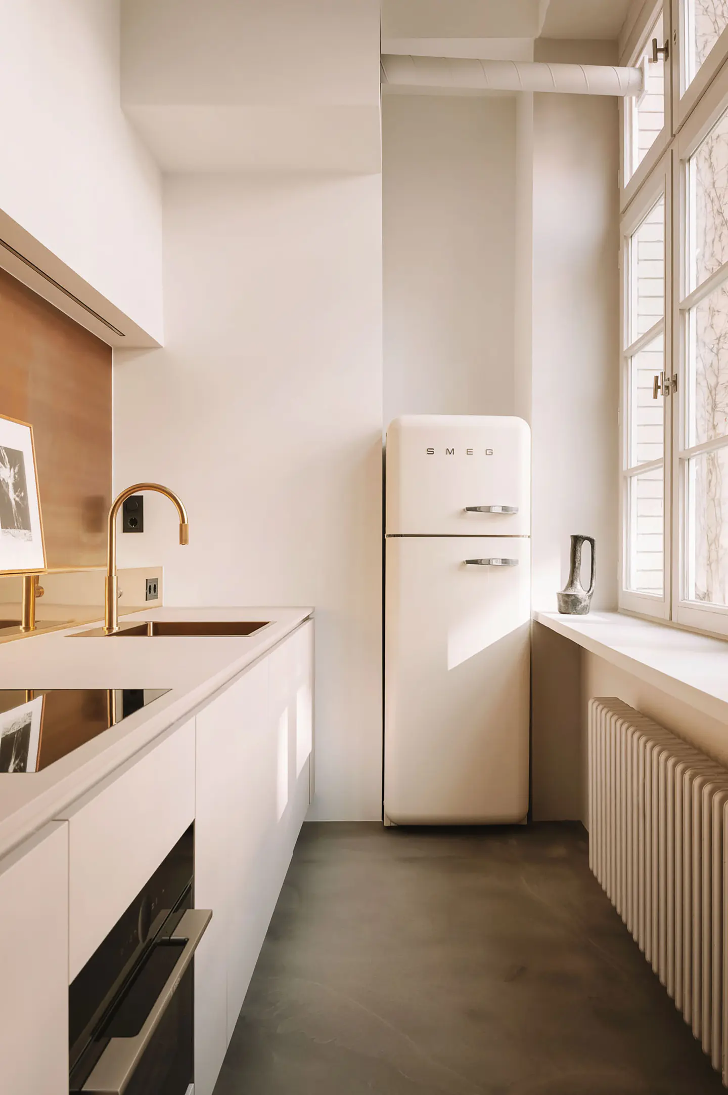 Modern minimalist kitchen with white cabinets, a cream Smeg refrigerator, gold faucet, and a window with a radiator.